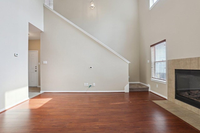 unfurnished living room featuring wood finished floors, a tile fireplace, baseboards, stairway, and a high ceiling