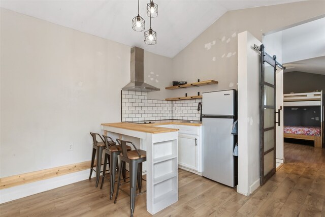 kitchen featuring butcher block countertops, freestanding refrigerator, a barn door, white cabinetry, and wall chimney exhaust hood