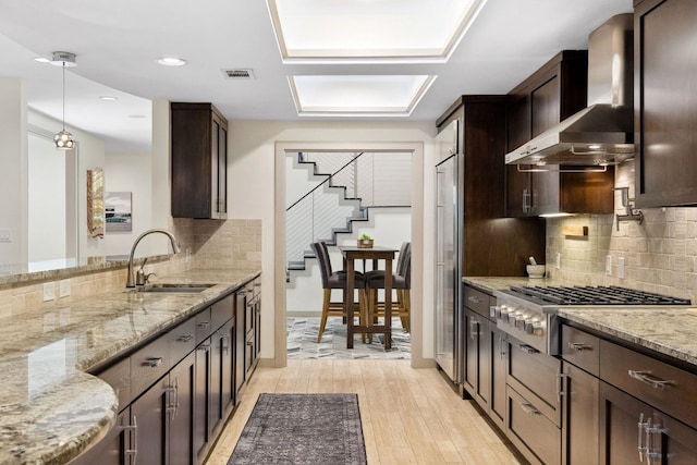 9 Swiftwater Trl, The Hills, TX 78738 - kitchen featuring wall chimney range hood, appliances with stainless steel finishes, dark brown cabinetry, light stone countertops, and light wood-style flooring
