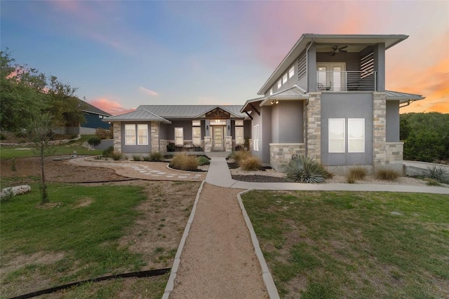 back of property at dusk with stucco siding, a yard, a ceiling fan, stone siding, and a balcony