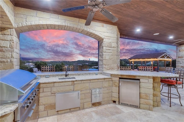 patio terrace at dusk featuring grilling area, a sink, an outdoor kitchen, outdoor wet bar, and a ceiling fan