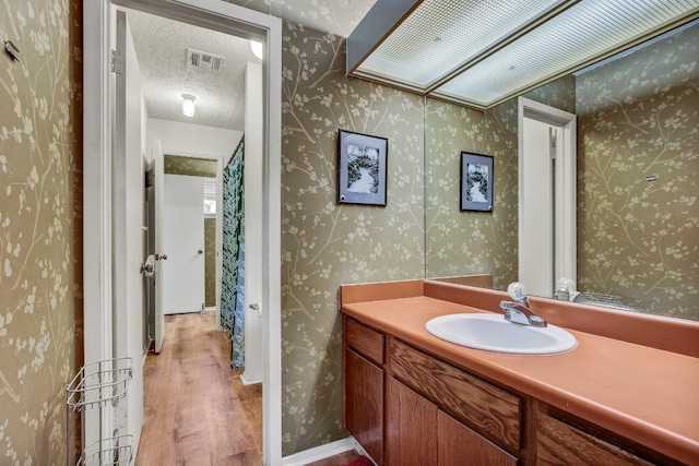 bathroom featuring a textured ceiling, visible vents, wallpapered walls, wood finished floors, and vanity