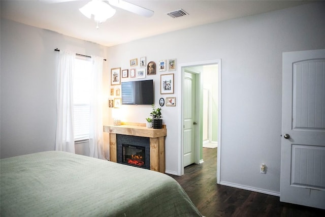 bedroom with baseboards, a glass covered fireplace, dark wood-style floors, visible vents, and a ceiling fan