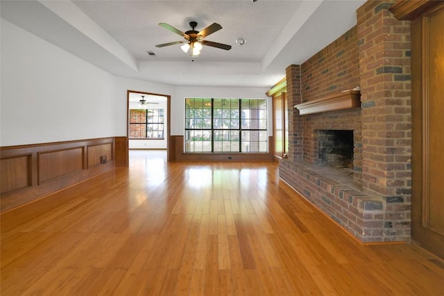 unfurnished living room with a fireplace, ceiling fan, a raised ceiling, light wood-type flooring, and wainscoting