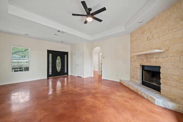 unfurnished living room featuring finished concrete flooring, a raised ceiling, ceiling fan, arched walkways, and a stone fireplace