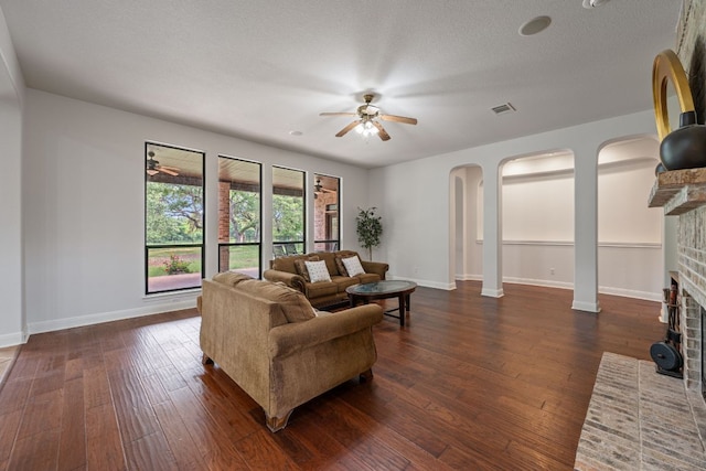 living room featuring ceiling fan, dark wood-style floors, baseboards, arched walkways, and a brick fireplace