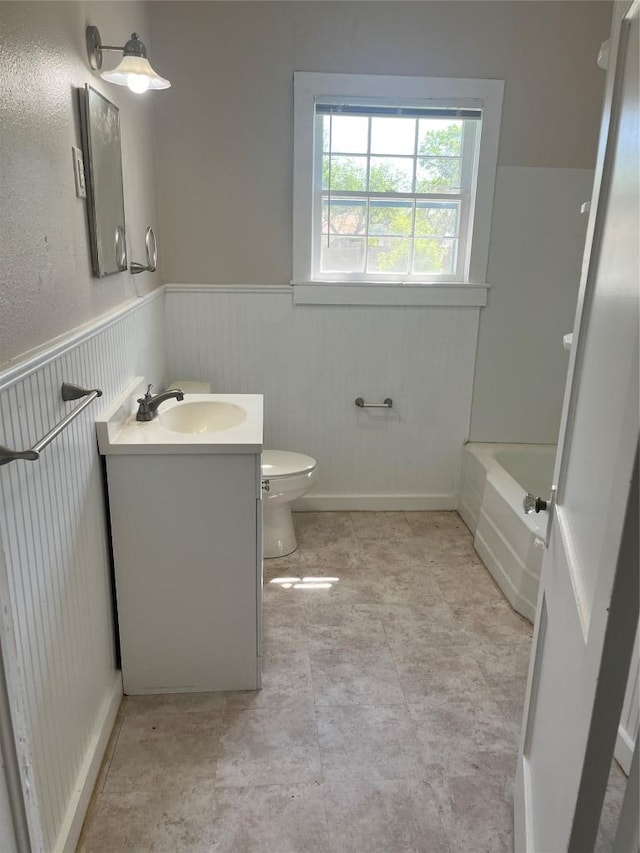 bathroom featuring toilet, a tub to relax in, vanity, and wainscoting
