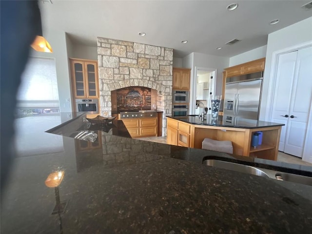 kitchen featuring visible vents, light brown cabinetry, dark stone counters, stainless steel appliances, and an island with sink