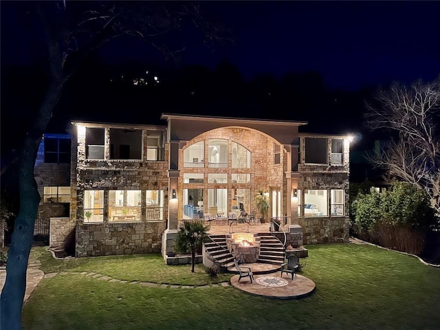 back of house at night with a lawn, stone siding, and a balcony