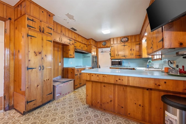 kitchen featuring stainless steel microwave, brown cabinets, light countertops, under cabinet range hood, and a peninsula