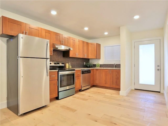 kitchen featuring brown cabinets, under cabinet range hood, decorative backsplash, light wood-style flooring, and appliances with stainless steel finishes