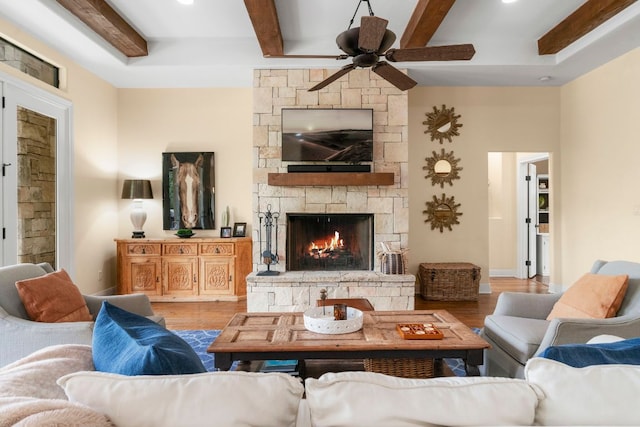 living room featuring beamed ceiling, light wood finished floors, a ceiling fan, and a stone fireplace