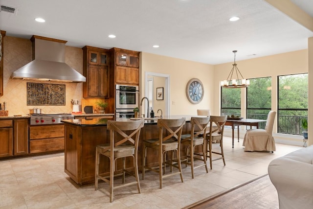 kitchen featuring wall chimney range hood, visible vents, stainless steel appliances, recessed lighting, and an island with sink