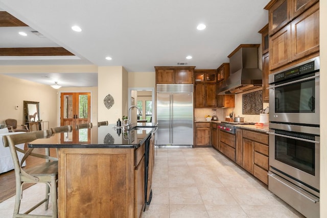kitchen featuring an island with sink, appliances with stainless steel finishes, a warming drawer, wall chimney exhaust hood, and a kitchen breakfast bar
