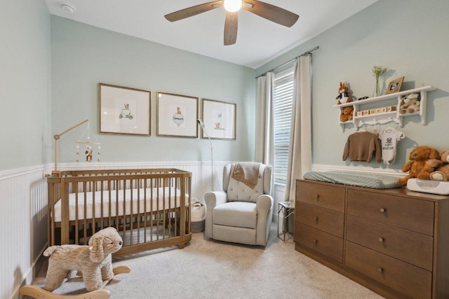 bedroom with a crib, light colored carpet, ceiling fan, and wainscoting