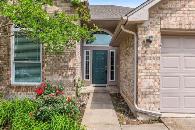 view of exterior entry with an attached garage, roof with shingles, and brick siding