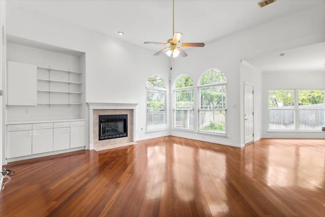 unfurnished living room with baseboards, built in shelves, hardwood / wood-style flooring, a tiled fireplace, and a ceiling fan