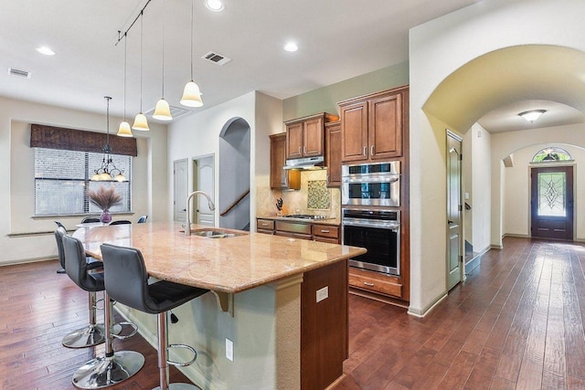 kitchen with a sink, arched walkways, under cabinet range hood, and light stone counters