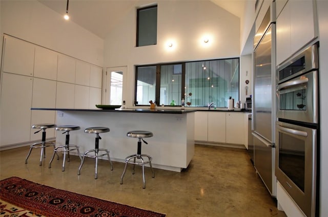 kitchen featuring a breakfast bar area, dark countertops, concrete flooring, stainless steel appliances, and white cabinetry