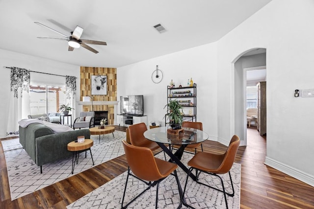 dining room featuring ceiling fan, a fireplace, arched walkways, wood finished floors, and visible vents