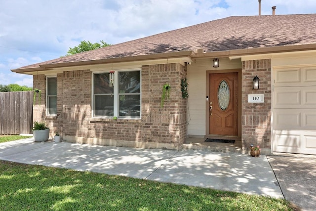 view of exterior entry featuring brick siding, a garage, roof with shingles, and fence