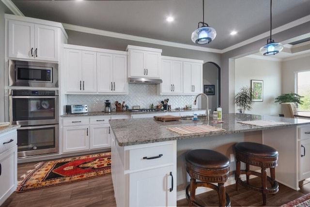 kitchen featuring crown molding, light stone countertops, white cabinetry, under cabinet range hood, and appliances with stainless steel finishes
