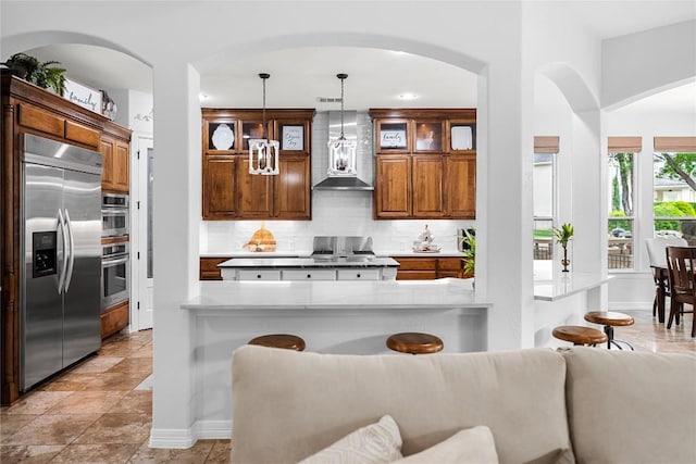 kitchen with wall chimney range hood, hanging light fixtures, built in fridge, backsplash, and glass insert cabinets
