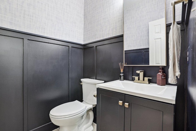 half bath featuring vanity, toilet, wainscoting, and a decorative wall