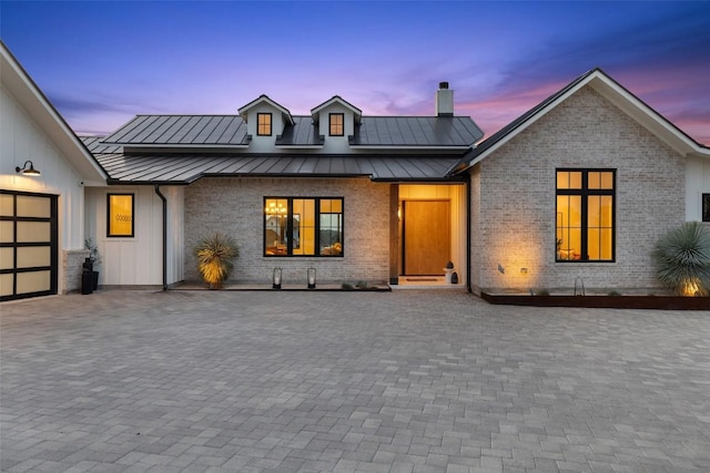 modern farmhouse style home featuring an attached garage, brick siding, a chimney, a standing seam roof, and metal roof