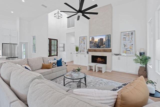 living area featuring visible vents, a high ceiling, a warm lit fireplace, a ceiling fan, and light wood-style flooring