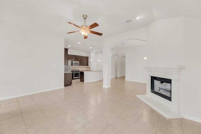 unfurnished living room featuring baseboards, ceiling fan, a fireplace with raised hearth, arched walkways, and light tile patterned floors