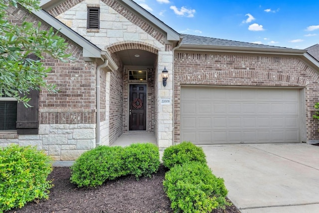 doorway to property featuring stone siding, roof with shingles, brick siding, concrete driveway, and an attached garage