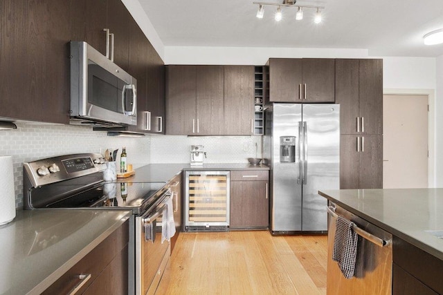 kitchen with dark brown cabinetry, beverage cooler, light wood-type flooring, tasteful backsplash, and stainless steel appliances