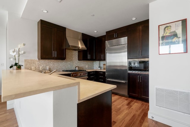 kitchen with wall chimney range hood, decorative backsplash, light countertops, a peninsula, and built in appliances