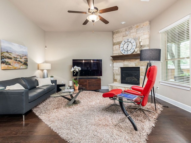 living area featuring a stone fireplace, a ceiling fan, baseboards, and wood finished floors