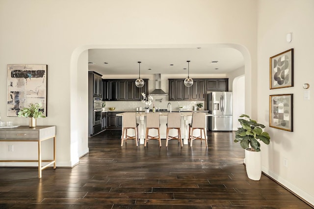 kitchen featuring appliances with stainless steel finishes, arched walkways, wall chimney exhaust hood, and an island with sink