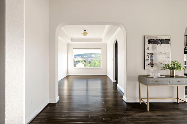 corridor featuring ornamental molding, baseboards, dark wood-style floors, a tray ceiling, and arched walkways