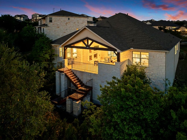 back of house at dusk with stairway, roof with shingles, and stone siding