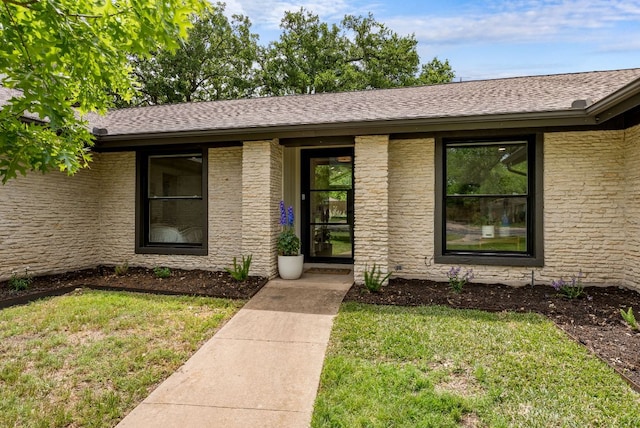 view of exterior entry featuring brick siding, roof with shingles, and a yard
