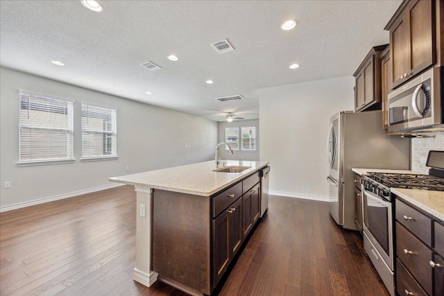 kitchen with a kitchen island with sink, appliances with stainless steel finishes, a sink, baseboards, and dark wood-style floors