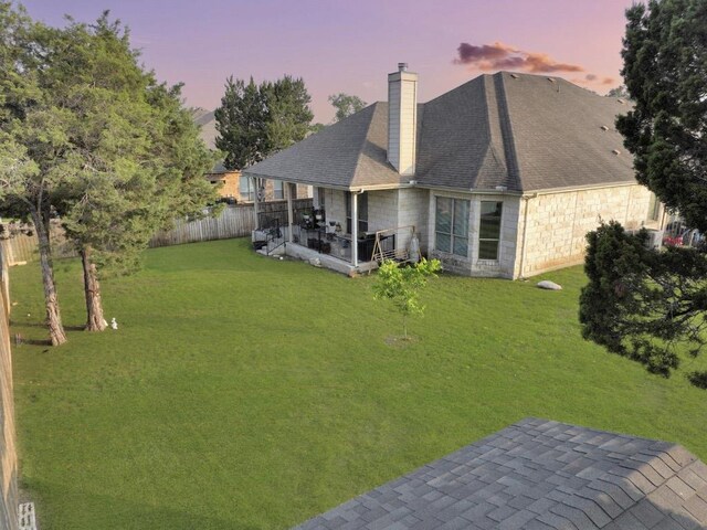 back of house at dusk with a lawn, fence, a chimney, and stone siding