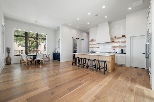 kitchen with a kitchen bar, open shelves, a barn door, white cabinets, and wall chimney exhaust hood