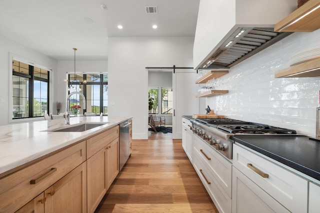 kitchen with wall chimney range hood, appliances with stainless steel finishes, open shelves, a sink, and light wood-style floors