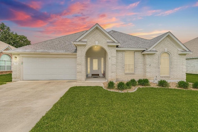 ranch-style home with a garage, driveway, roof with shingles, brick siding, and a yard