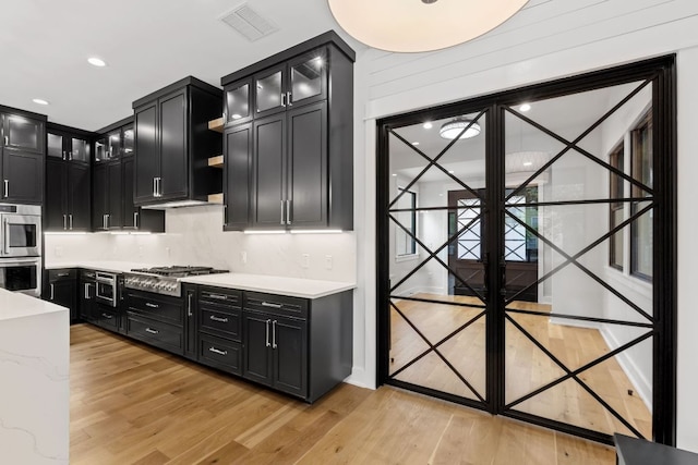 kitchen with light wood-style floors, dark cabinets, stainless steel appliances, visible vents, and glass insert cabinets
