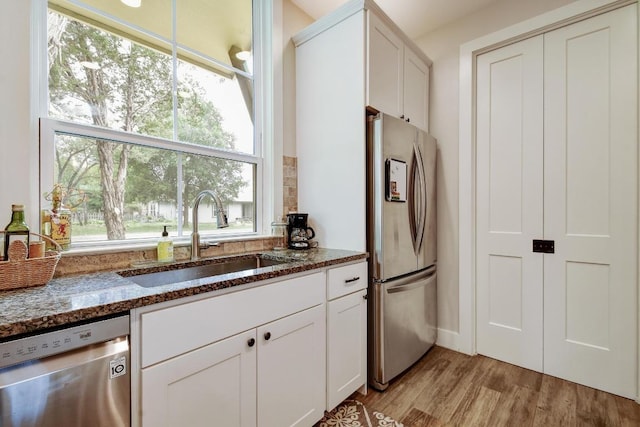 kitchen featuring appliances with stainless steel finishes, a sink, light wood finished floors, dark stone countertops, and a wealth of natural light