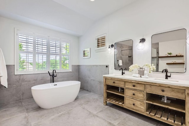 bathroom with a sink, double vanity, tile walls, vaulted ceiling, and a soaking tub