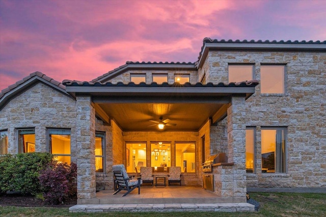 back of house at dusk featuring a patio, ceiling fan, a tiled roof, and stone siding