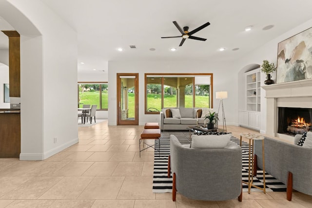 tiled living room featuring ceiling fan, arched walkways, a lit fireplace, baseboards, and visible vents