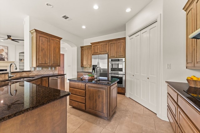 kitchen featuring a kitchen island with sink, a sink, dark stone countertops, appliances with stainless steel finishes, and backsplash
